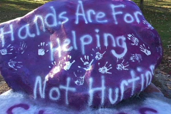 Large rock painted purple with hand prints for Relationship Abuse Awareness Month. Saying "Hands are for helping not hurting"